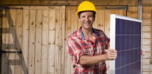 Portrait of smiling engineer holding solar panel in front of cabin