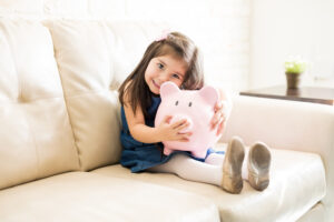 Portrait of beautiful little girl holding a big piggy bank in hands while sitting on sofa in living room
