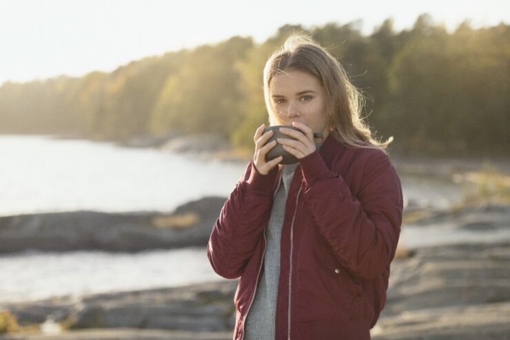 Tüdruk looduses, seisab järve kaldal, joob kuuma jooki.