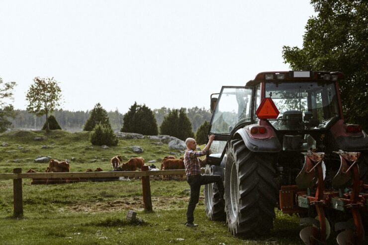 Pildil on mees farmis, kes ronib oma traktorisse. Taustal söövad lehmad muru ning on kaugelt näha ka metsapiiri.